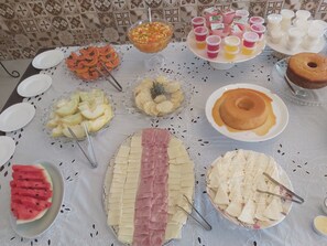 Mesa de buffet com diversos alimentos, incluindo frutas, queijos e sobremesas.