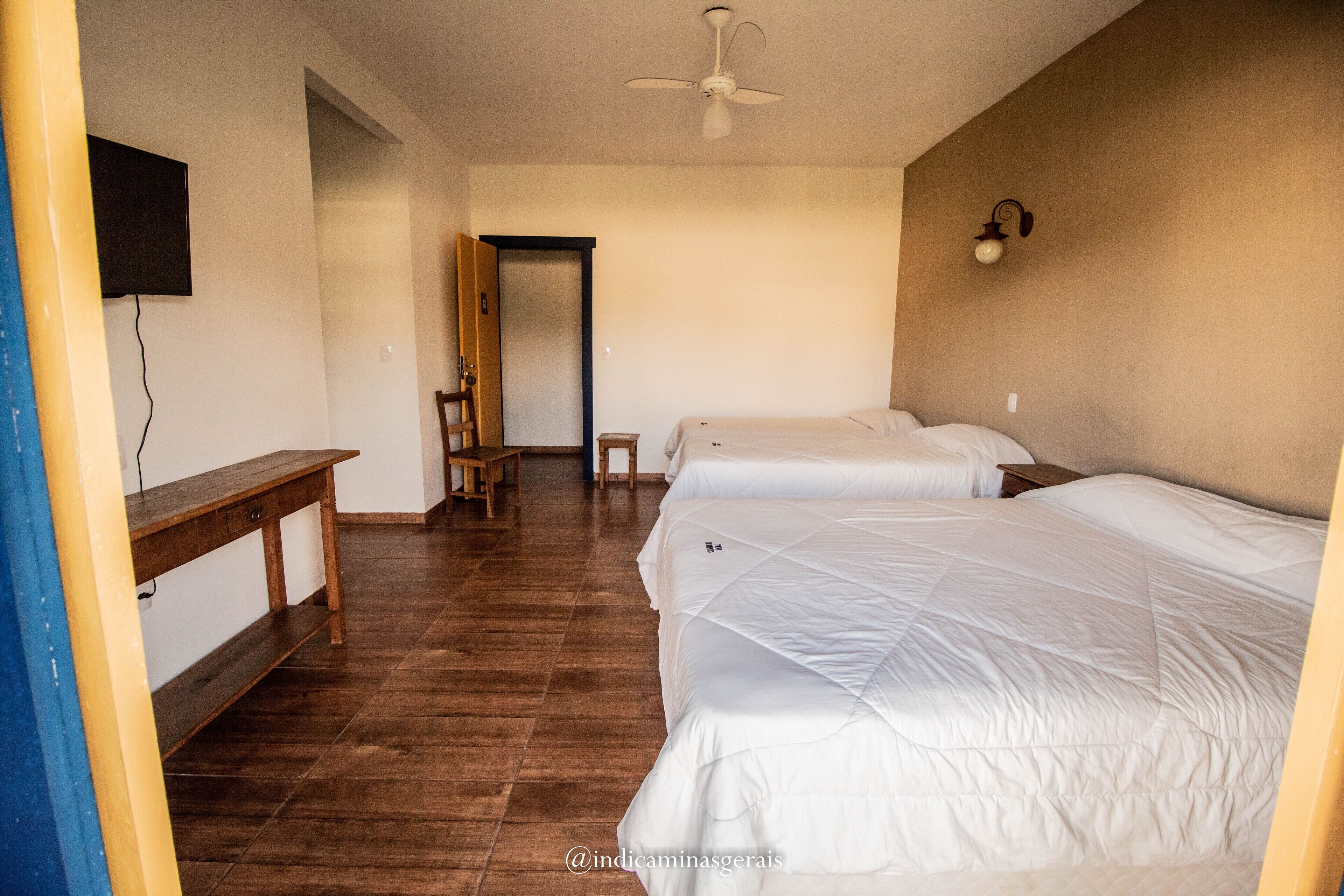 A hotel room with a wooden floor, a bed with white bedding, a desk, a chair, and a ceiling fan.