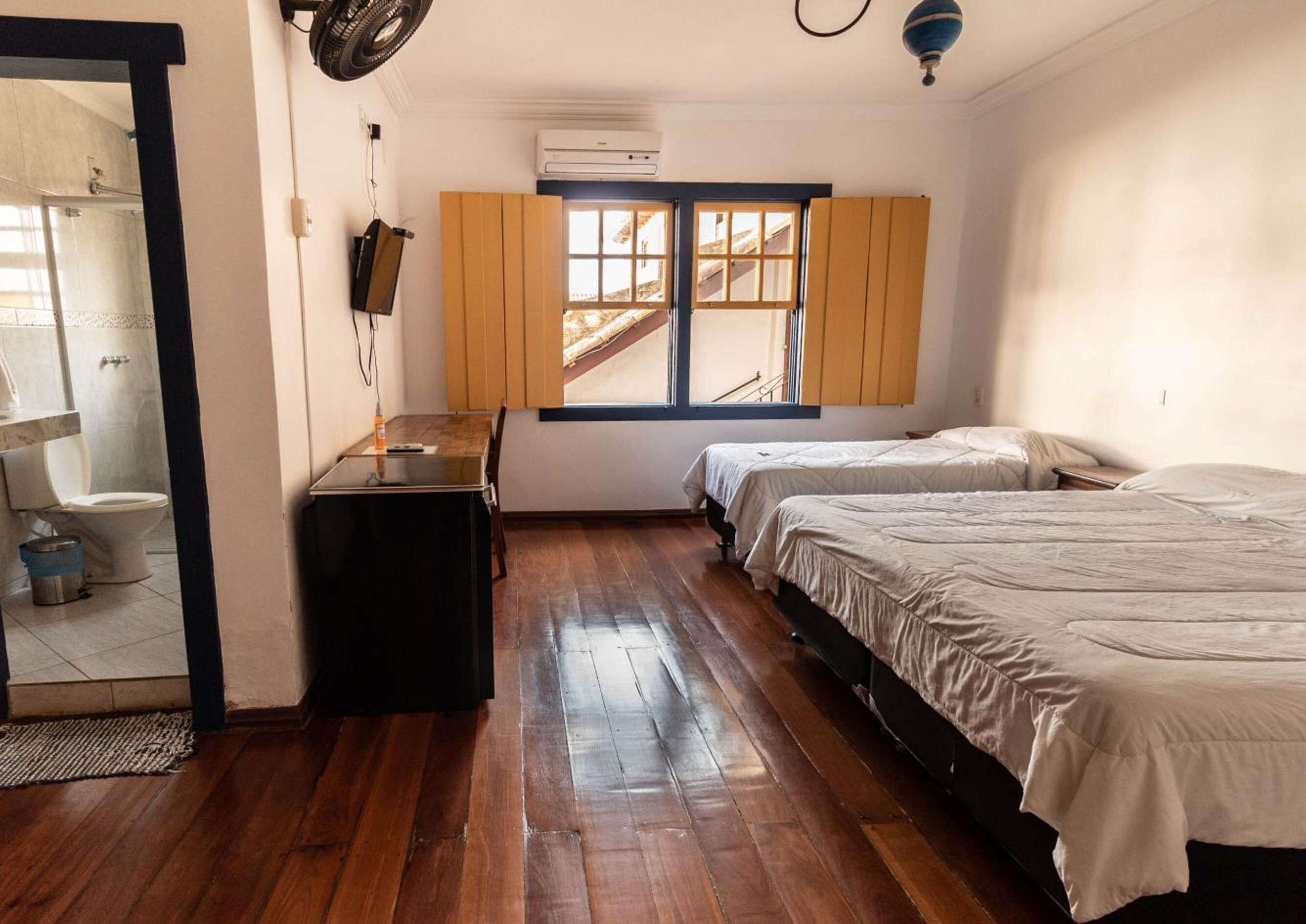 A compact room with a wooden floor, a desk, two beds, a window with wooden shutters, and an air conditioning unit.