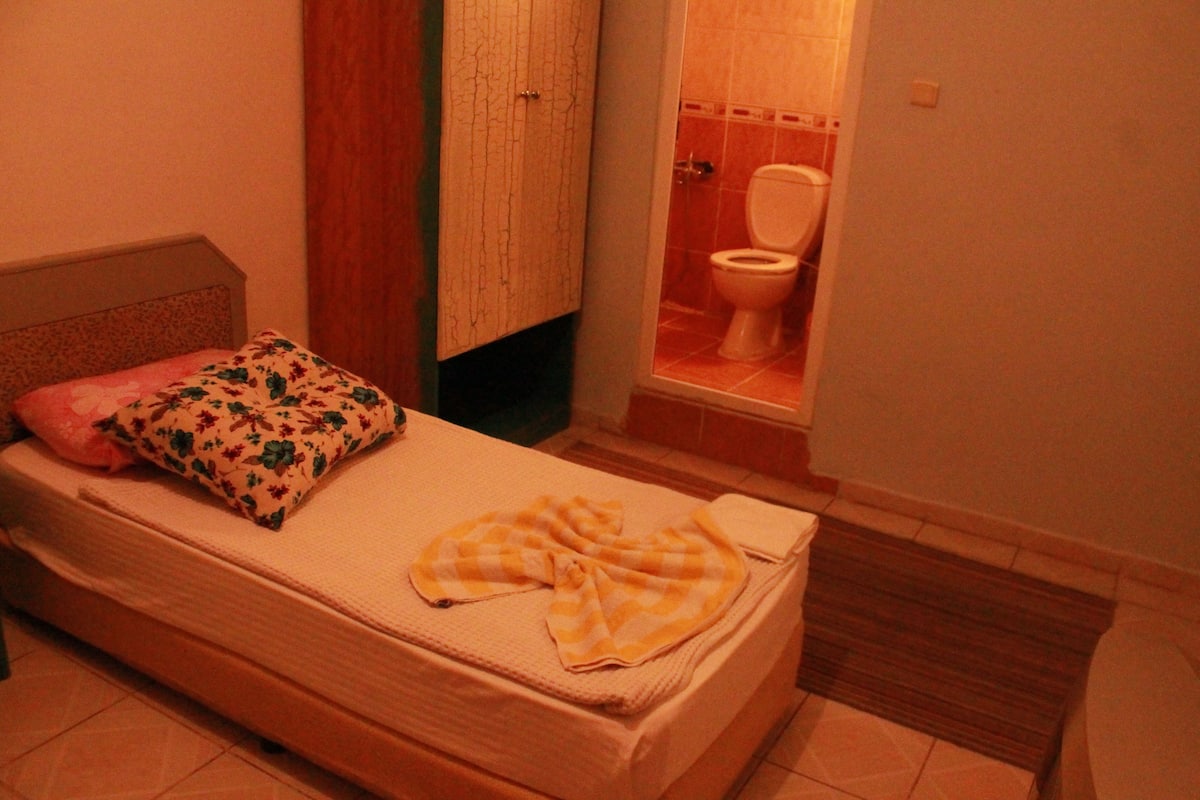 A small bedroom with a single bed, a wooden wardrobe, and a bathroom visible through an open door.
