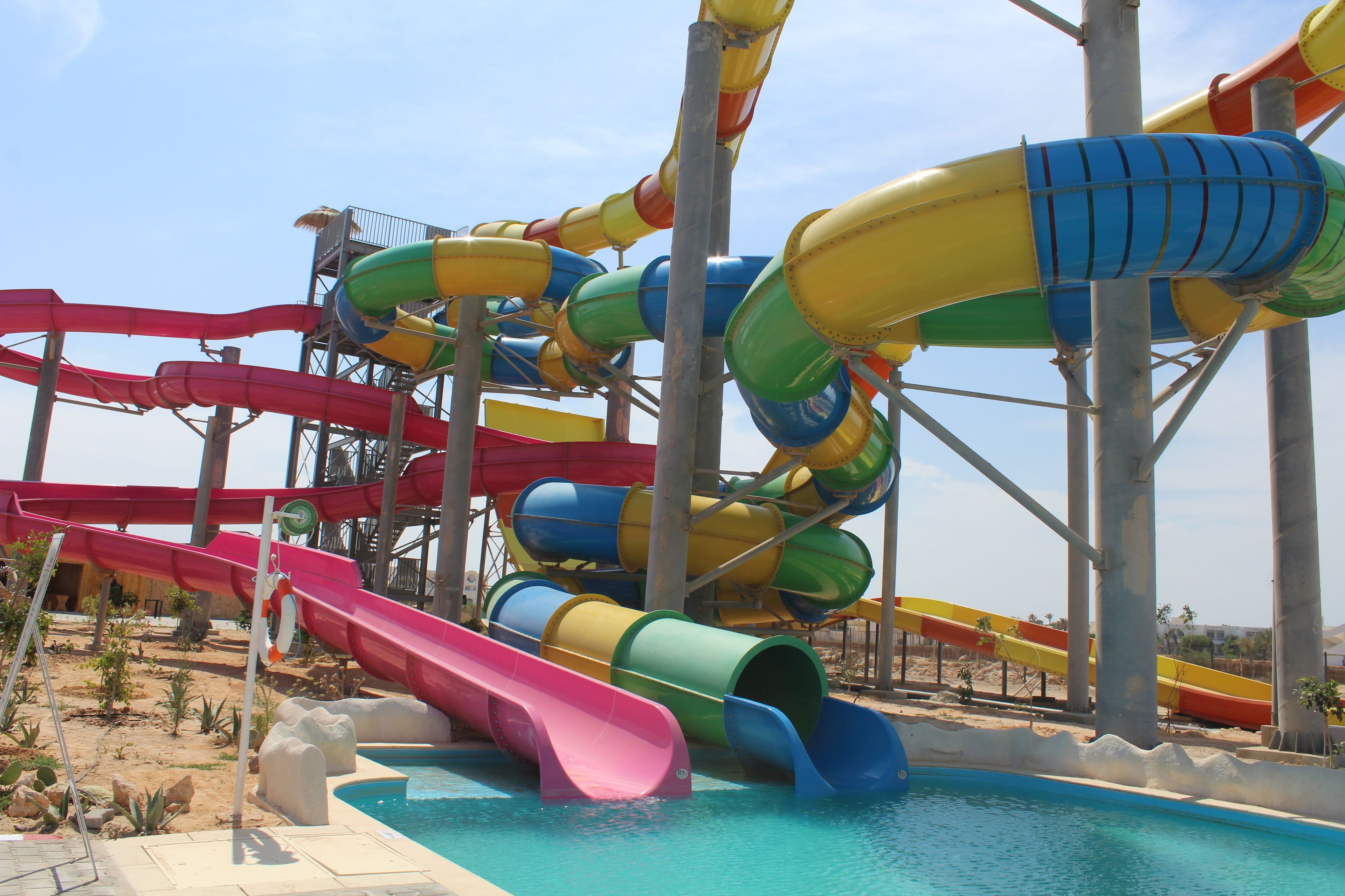 Um parque aquático com várias escorregas em diferentes cores, uma piscina e uma área arenosa com plantas.