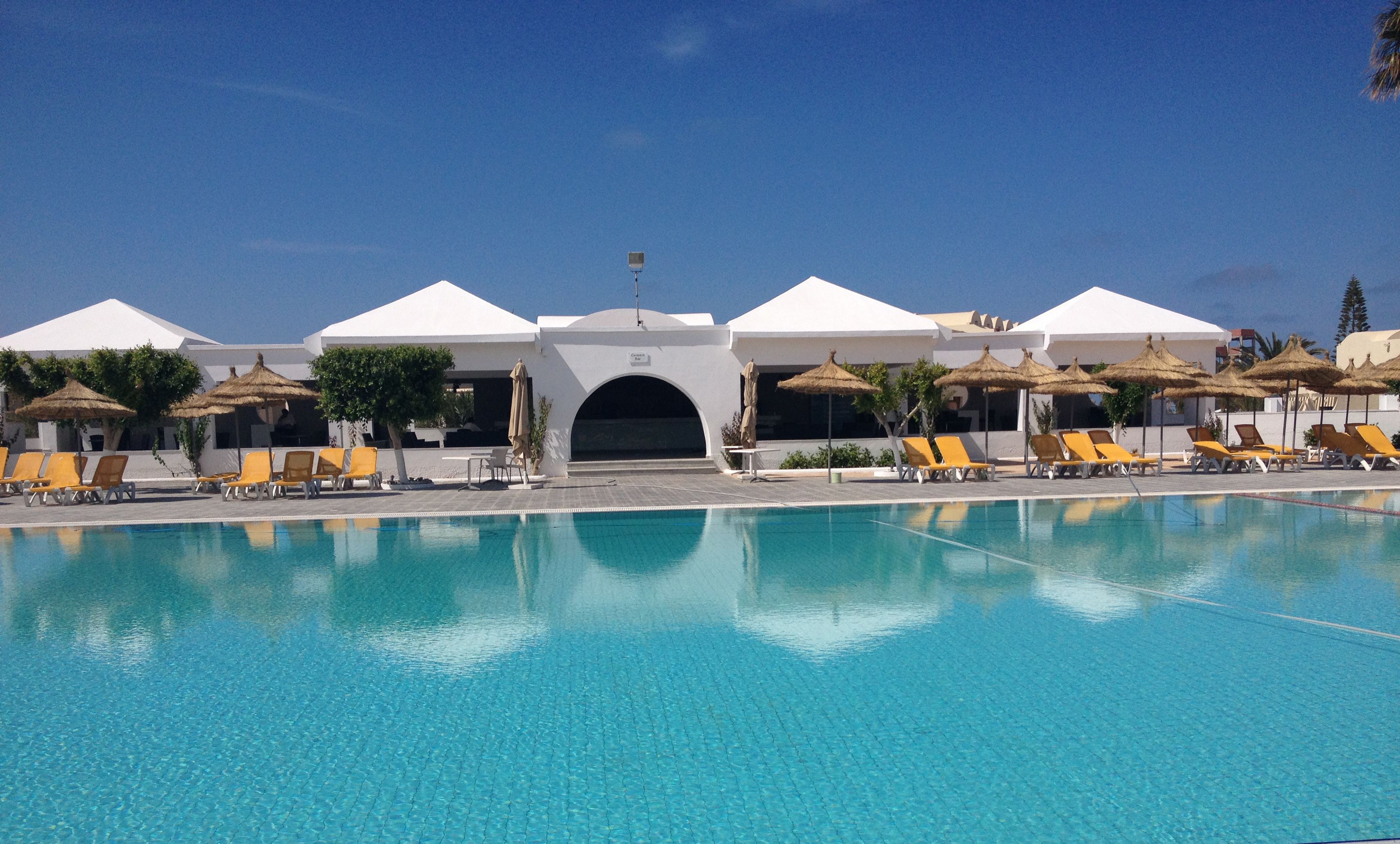 Une piscine entourée de chaises longues et de parasols, elle-même bordée de bâtiments blancs aux portes en arc de cercle.