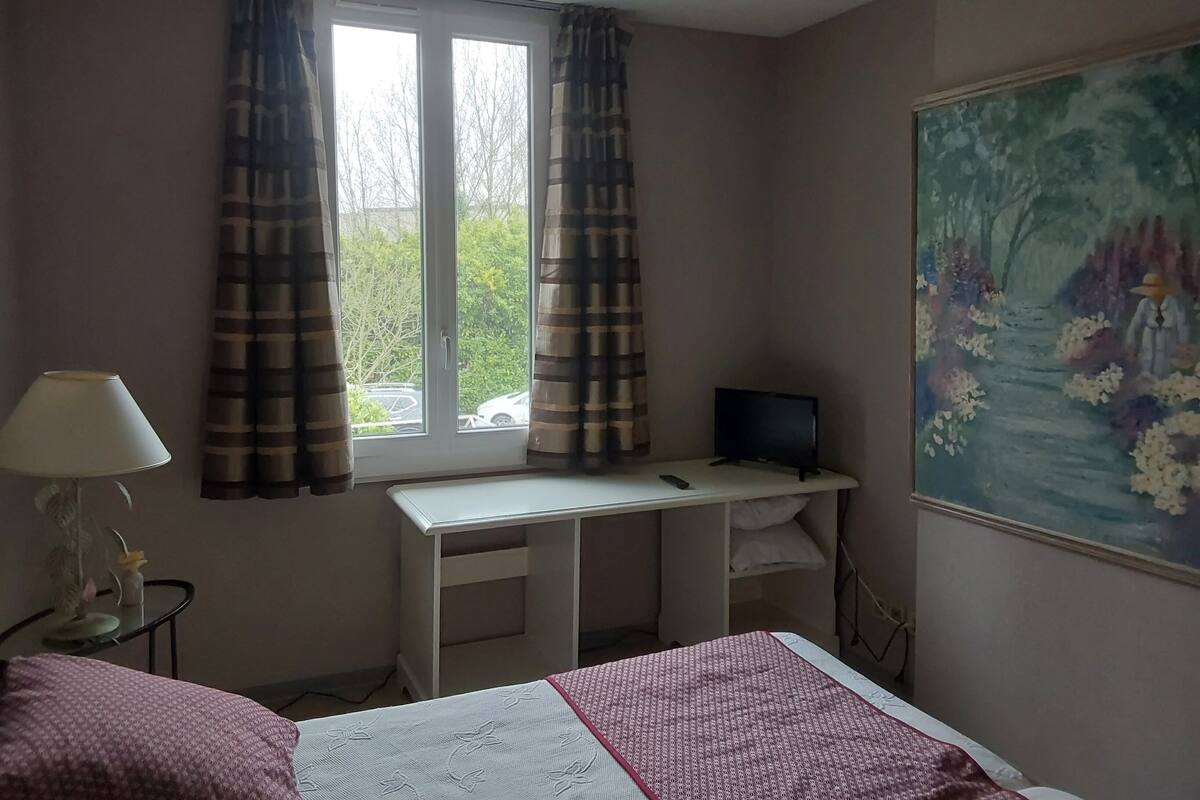 Un dormitorio con cama, escritorio con computadora, lámpara y un cuadro en la pared.