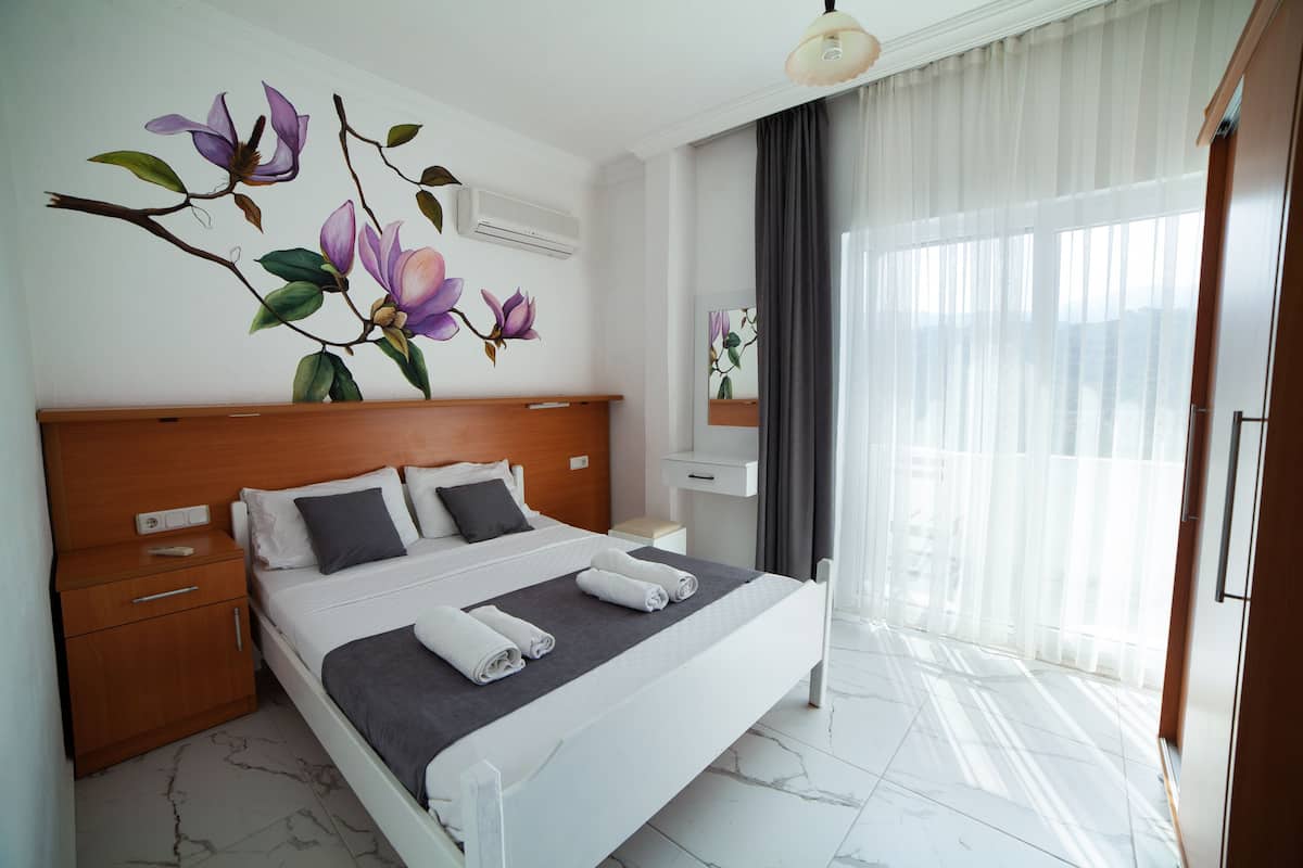 Quarto de hotel moderno com uma cama grande, cabeceira de madeira, mesa de cabeceira e uma janela ampla com cortinas leves.