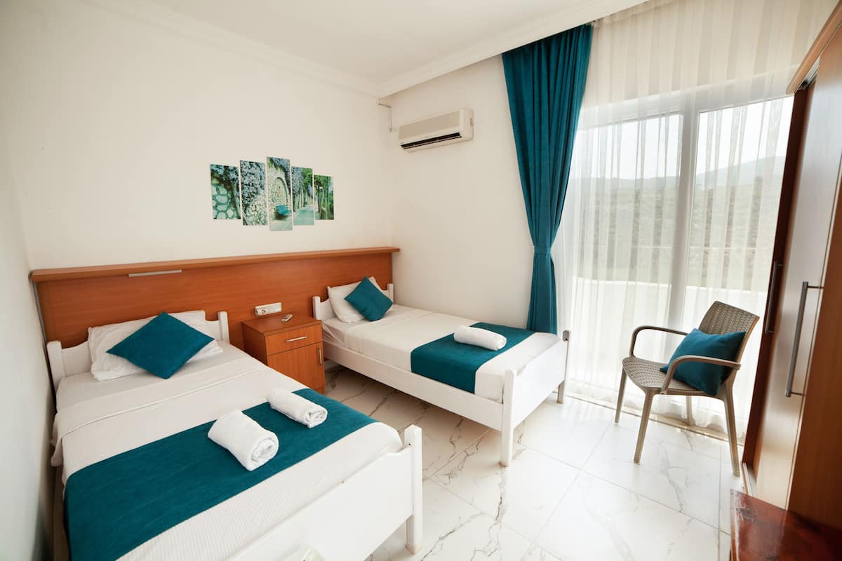 Quarto de hotel com duas camas de solteiro, cabeceira de madeira, uma mesa pequena, uma cadeira e uma janela com cortinas.
