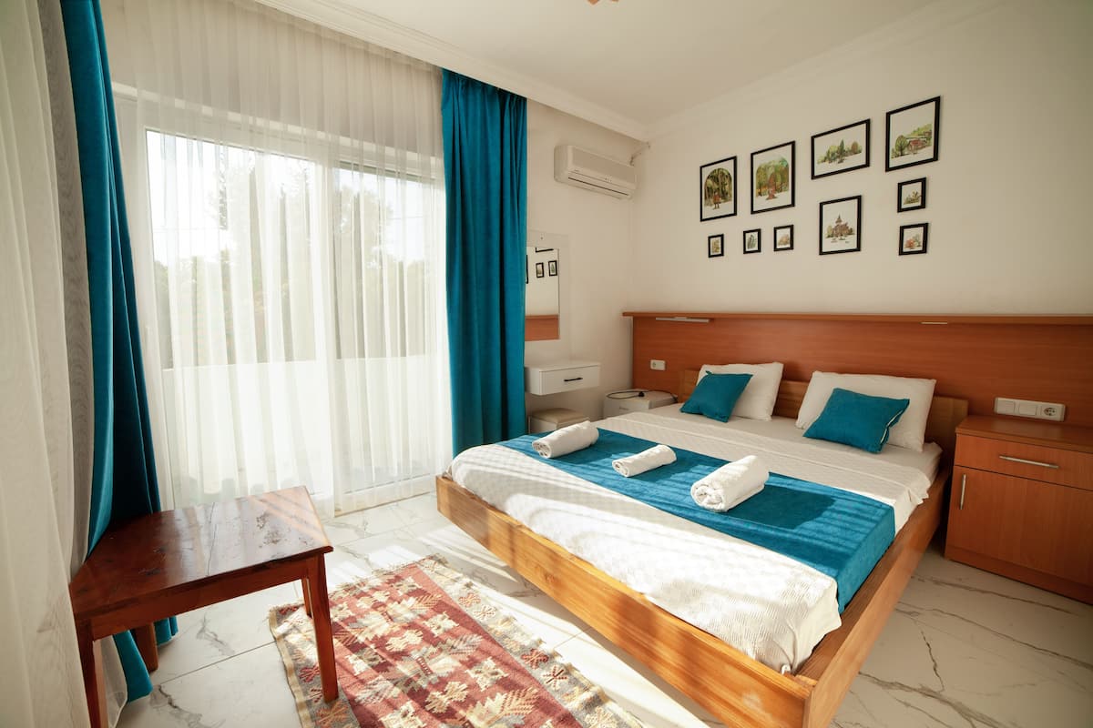 Quarto de hotel com uma cama de madeira, uma mesinha, uma escrivaninha e uma parede com quadros emoldurados.