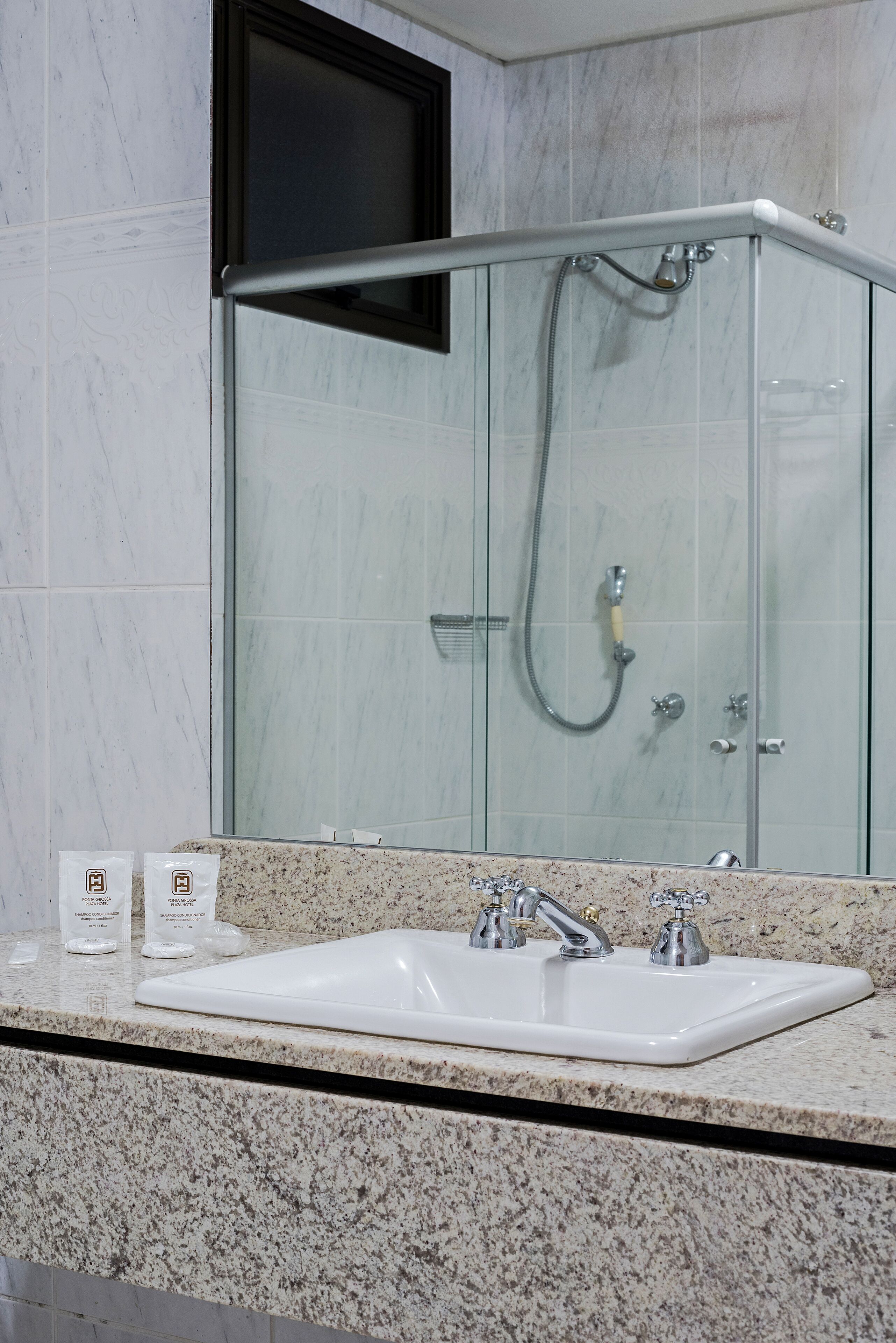 A bathroom with a glass-enclosed shower, a white sink with two faucets, and a granite countertop.
