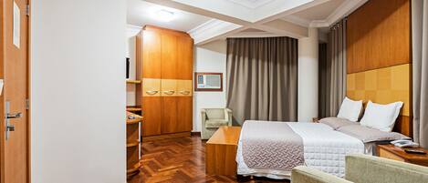 Habitación de hotel con piso de madera, cama con colcha blanca y beige, armario de madera, escritorio, silla y mesita de noche con control remoto.