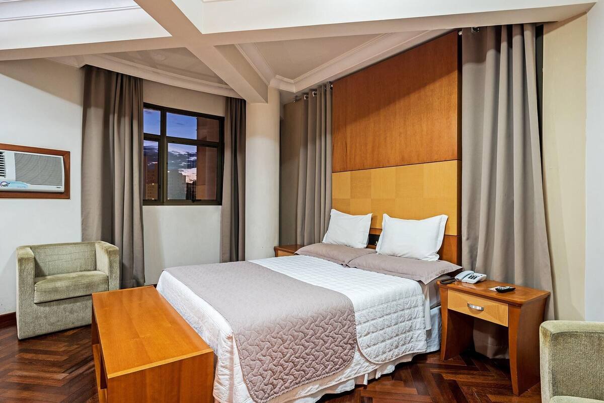 A hotel room with a large bed, a wooden nightstand, a desk, a chair, and a window with curtains.