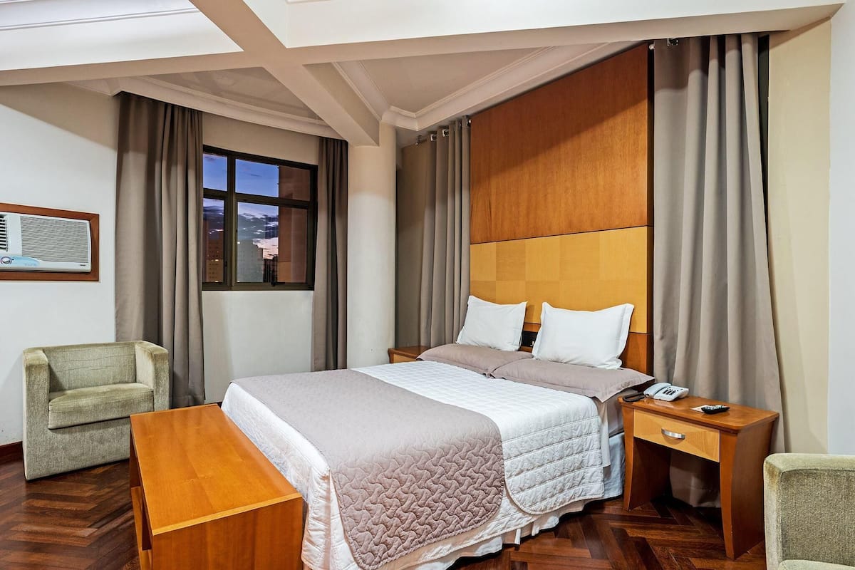 A hotel room with a large bed, a wooden nightstand, a desk, a chair, and a window with curtains.