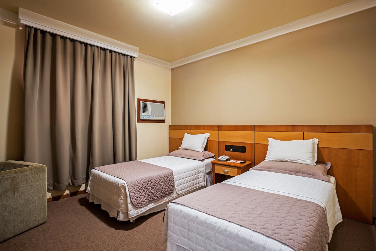 A hotel room with two beds, a bedside table with a phone, a small chair, and a window with curtains.