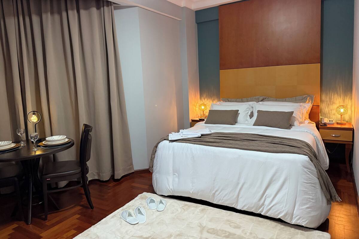 A hotel room with a bed, bedside tables with lamps, a dining table with chairs, and a window with curtains.