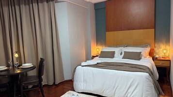 A hotel room with a bed, bedside tables with lamps, a dining table with chairs, and a window with curtains.