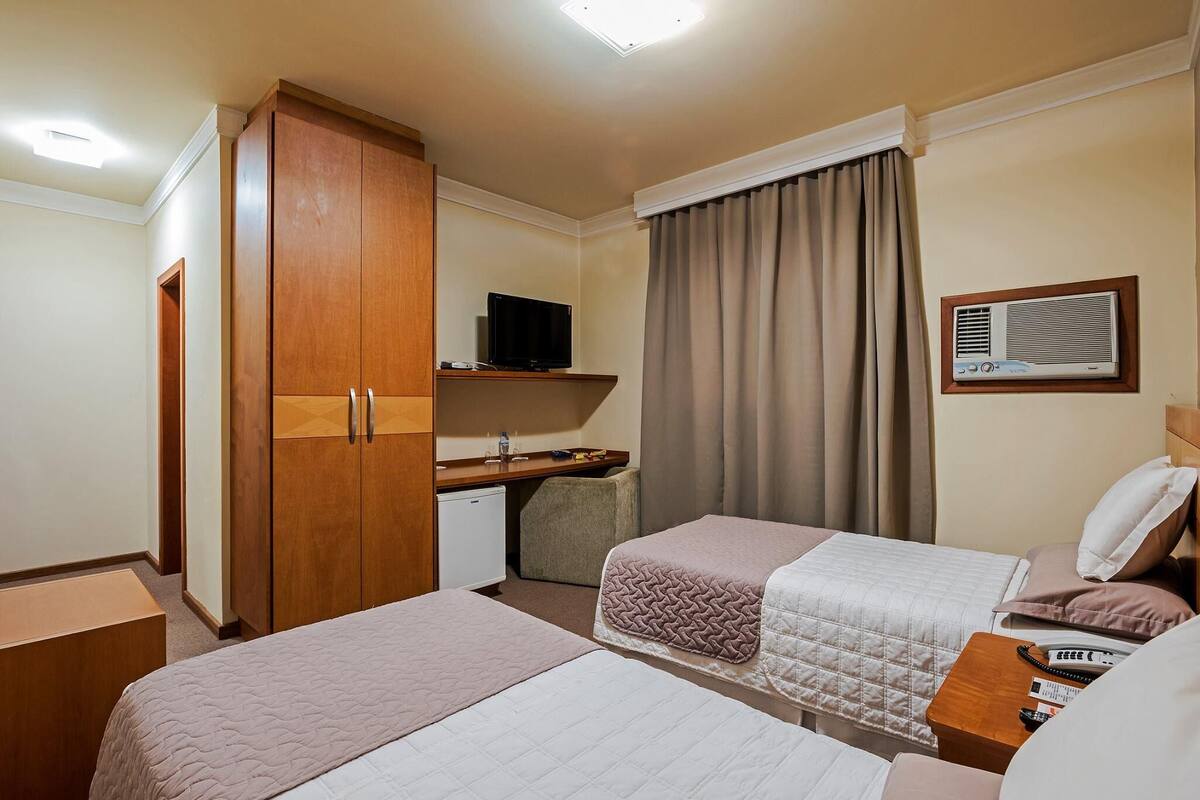 A hotel room with two beds, a desk with a television, a wardrobe, and an air conditioning unit.