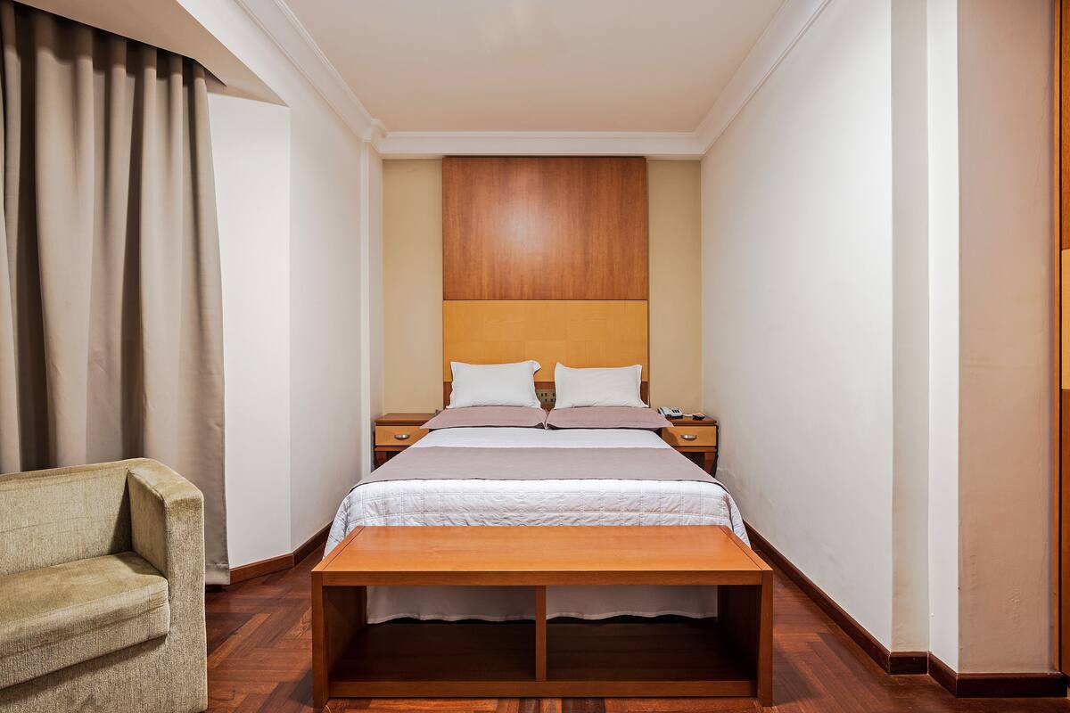 A hotel room with a bed, bedside tables, a wooden bench, a chair, and a large window with curtains.