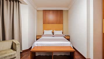 Een hotelkamer met een bed, nachtkastjes, een houten bankje, een stoel en een groot raam met gordijnen.