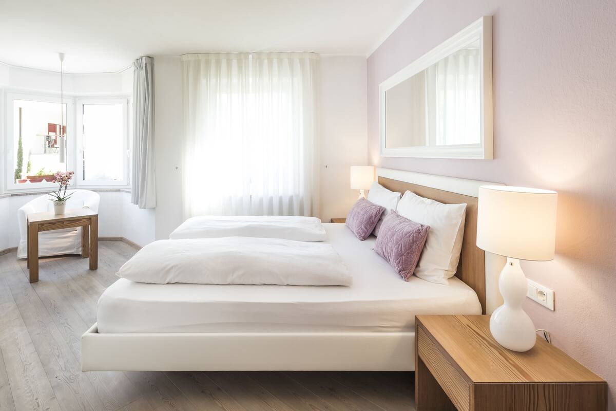 A modern hotel room with a large bed, bedside tables, a small desk, and a window with curtains.