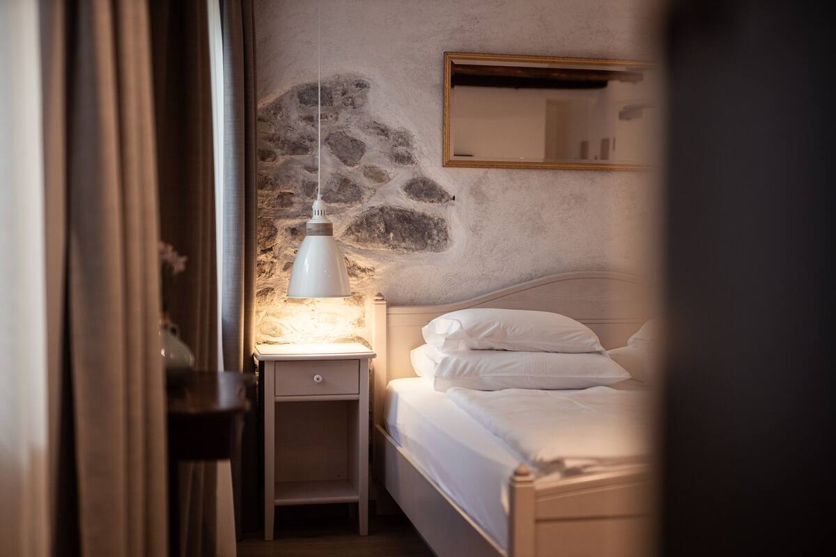 A bedroom with a stone wall, a bedside lamp, a bed with white bedding, and a mirror above the bed.