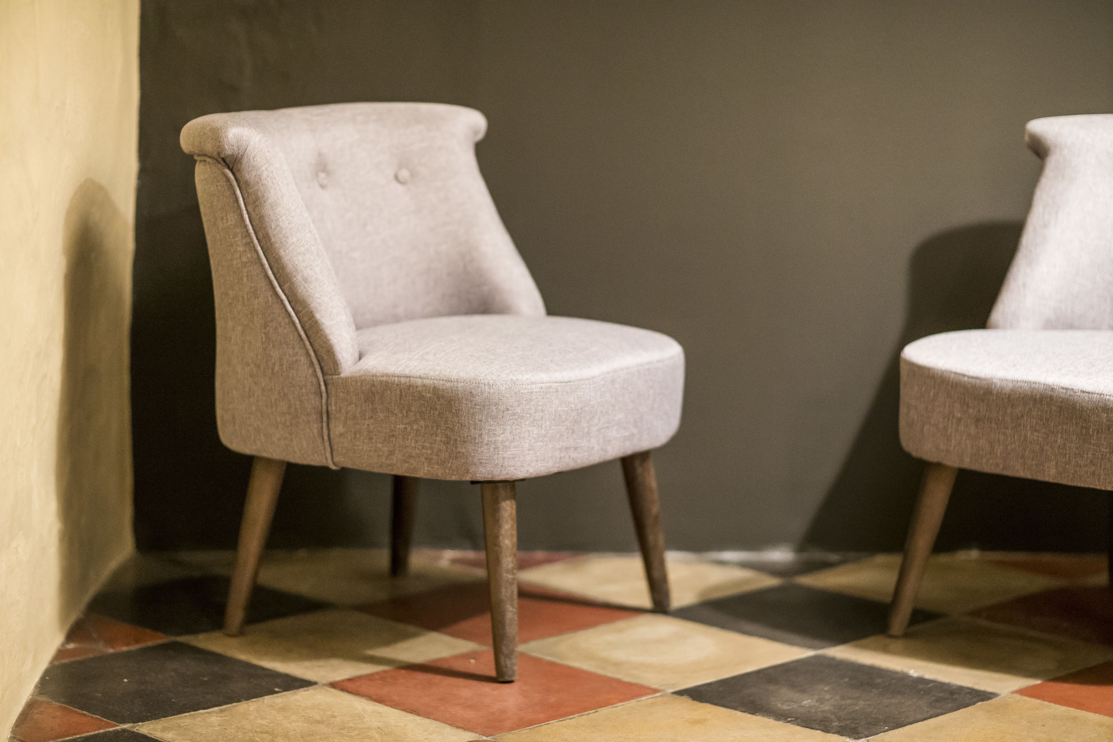 Two light-colored armchairs with wooden legs, positioned against a wall on a checkered floor.