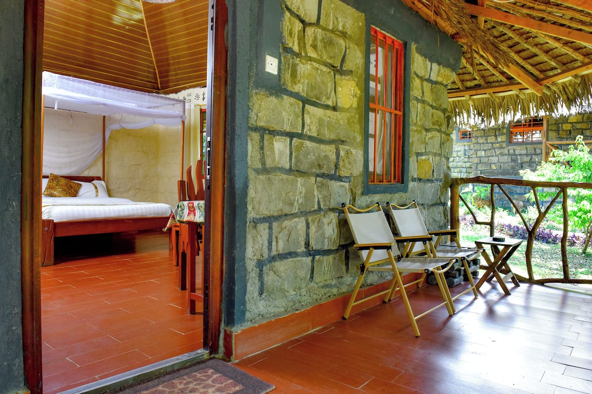 A room with a bed, a small table, and a chair. There is a stone wall and a wooden ceiling.