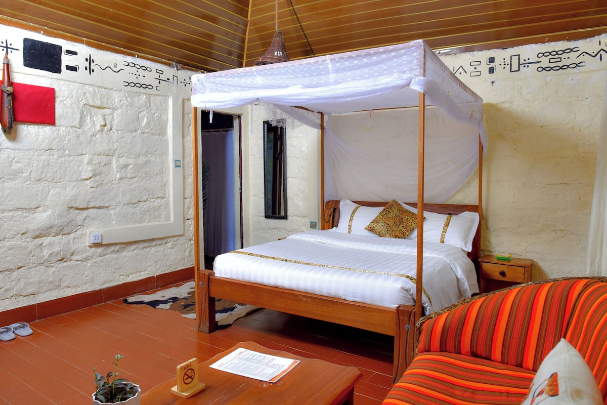 A four-poster bed with a canopy, a wooden nightstand, a striped armchair, and a small table with a plant and a menu.