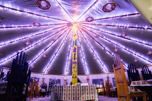 Una carpa con un poste central, rodeada de mesas y sillas, e iluminada con luces colgantes.