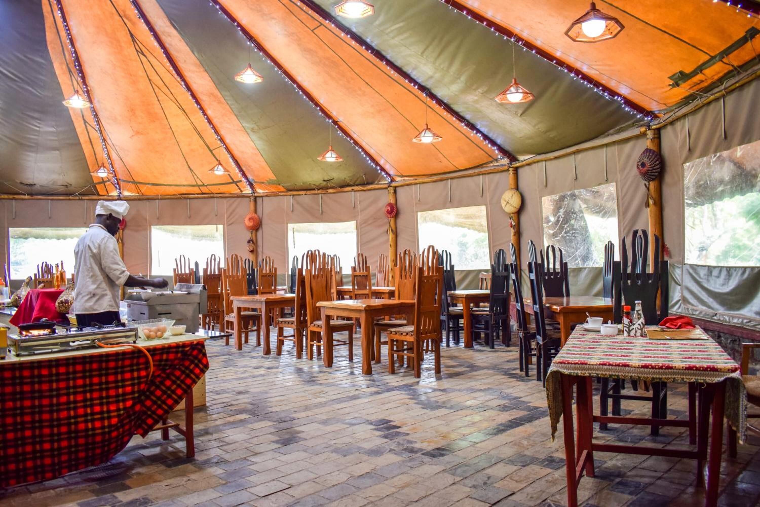 Un área de comedor con carpas, mesas y sillas, un chef preparando comida, y ventanas grandes.