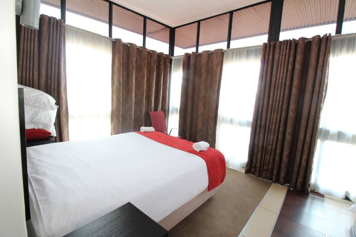 A hotel room with a large bed, a red pillow, a white pillow, and a red blanket. There is a window with brown curtains and a view of the outside.