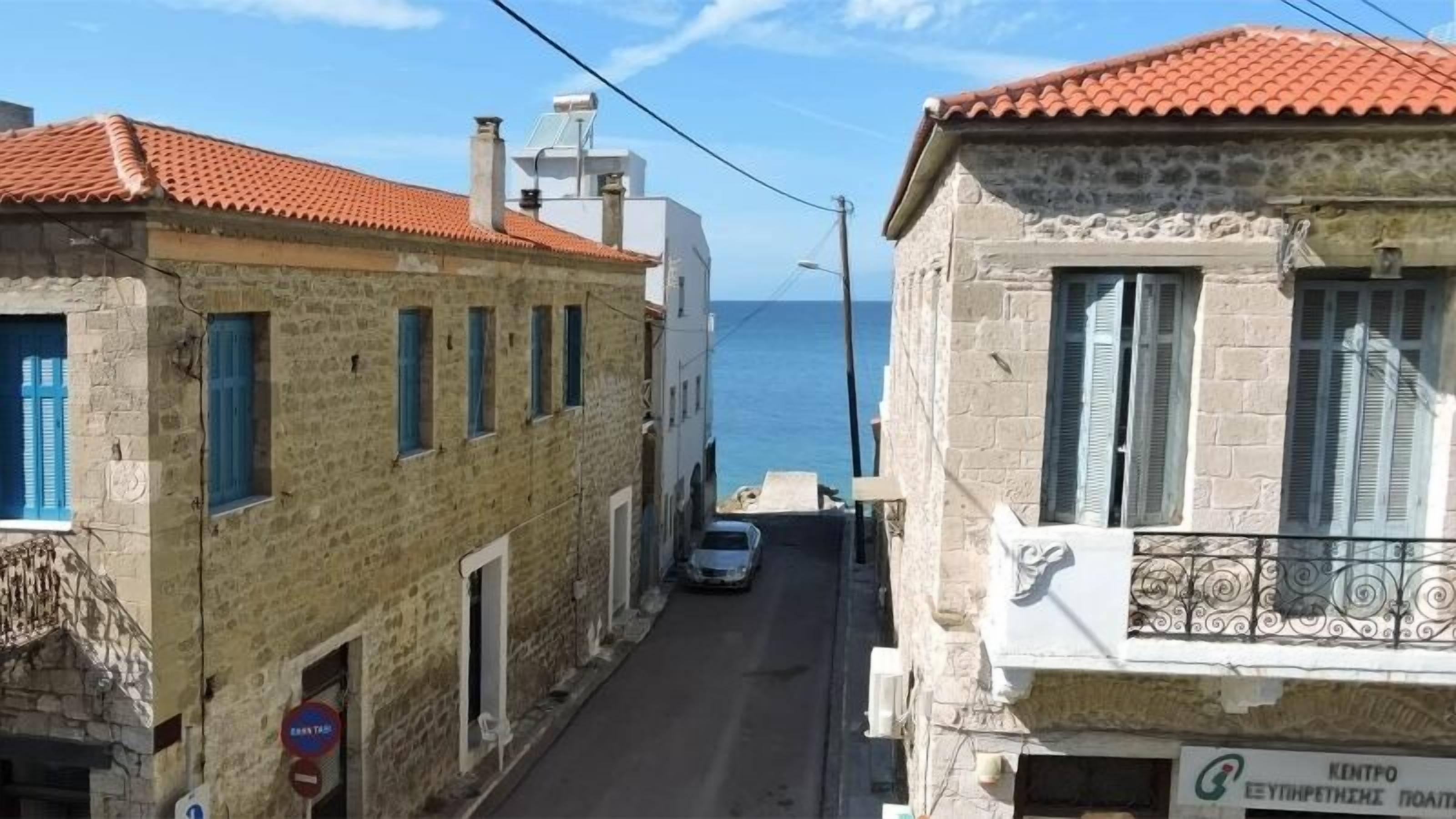 Una calle estrecha con edificios de piedra, un coche estacionado al costado y vista al mar en la distancia.
