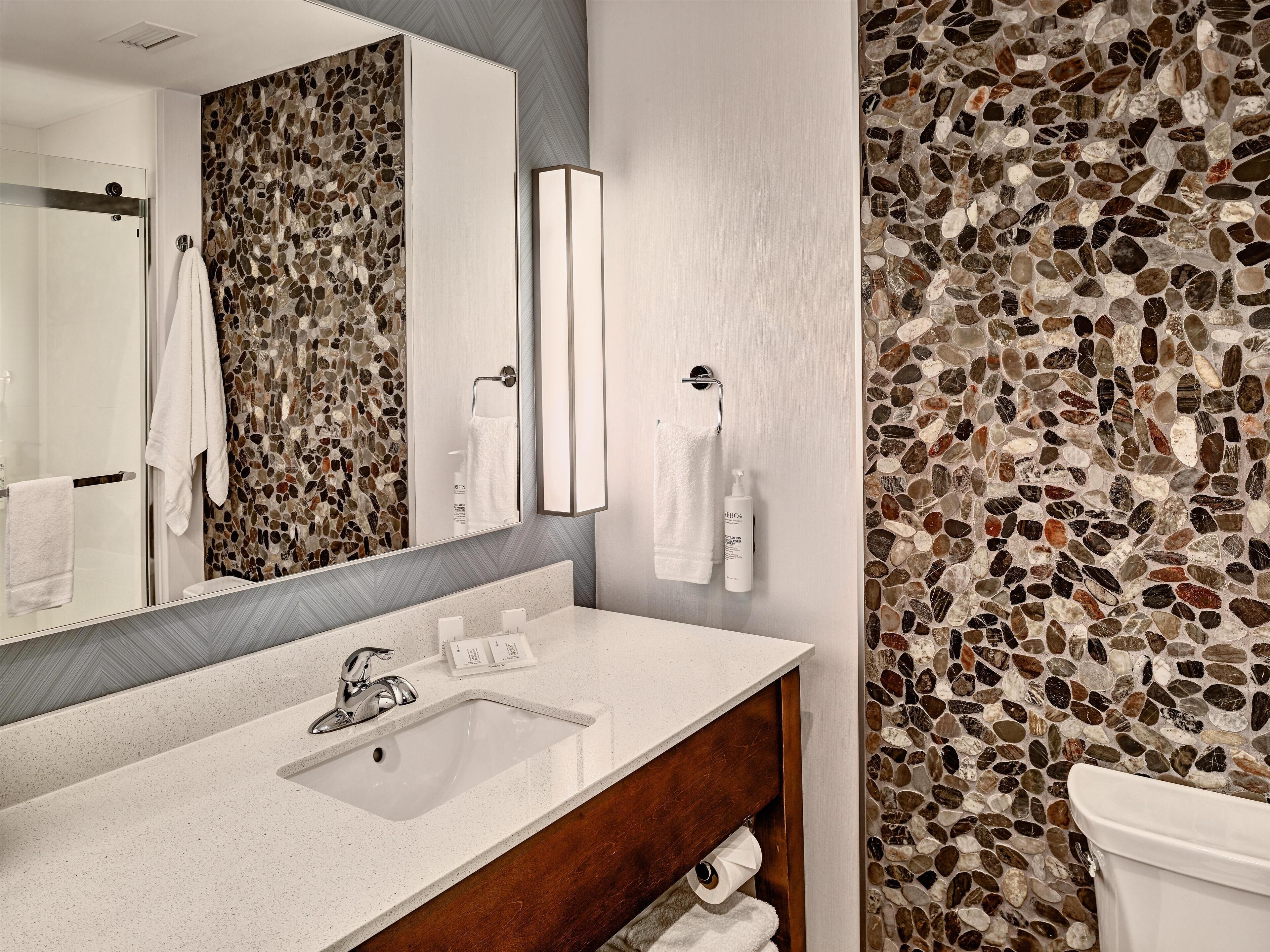 Un baño moderno con pared de mosaico de piedra, un espejo grande, un lavamanos blanco y un mueble de tocador de madera.