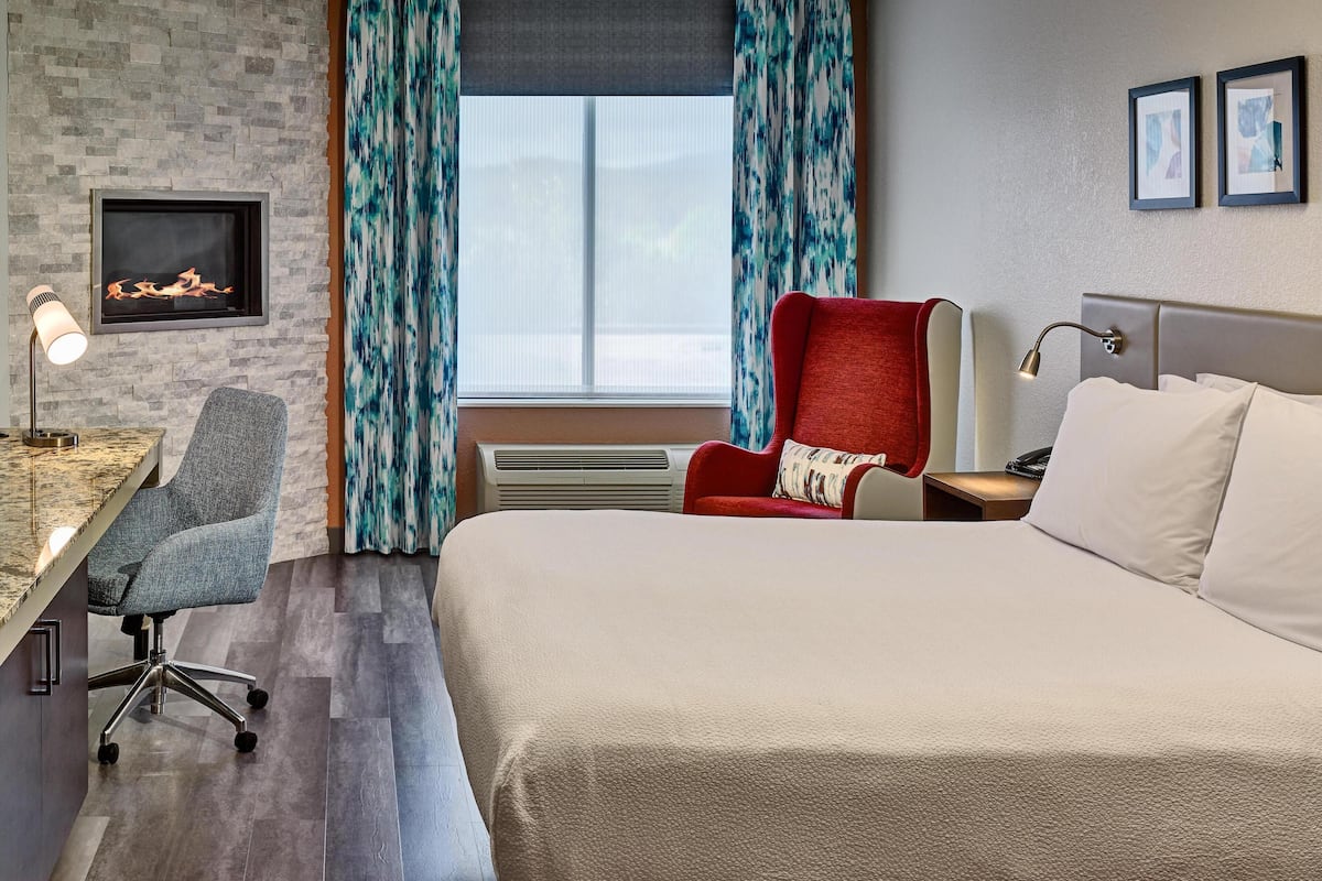 A hotel room with a bed, a desk with a chair, a red armchair, a fireplace, and curtains.
