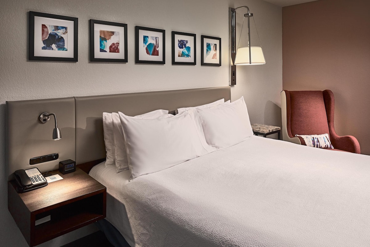A hotel room with a large bed, a bedside table with a phone and a digital clock, a chair, and wall-mounted artwork.