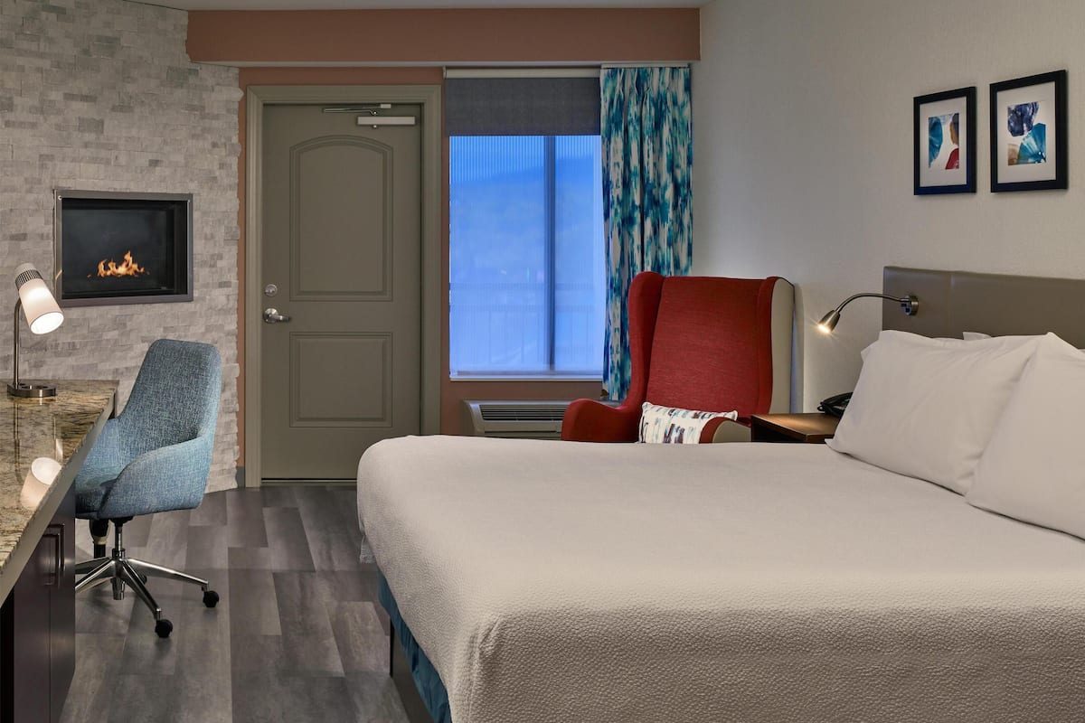 A hotel room with a bed, a desk with a chair, a fireplace, a window with curtains, and two framed pictures on the wall.