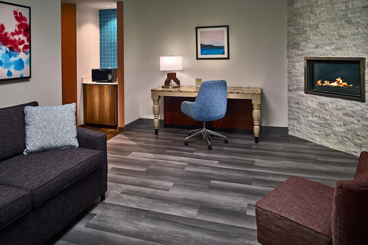 A modern hotel room with a dark gray sofa, a blue office chair, a wooden desk, and a stone accent wall.