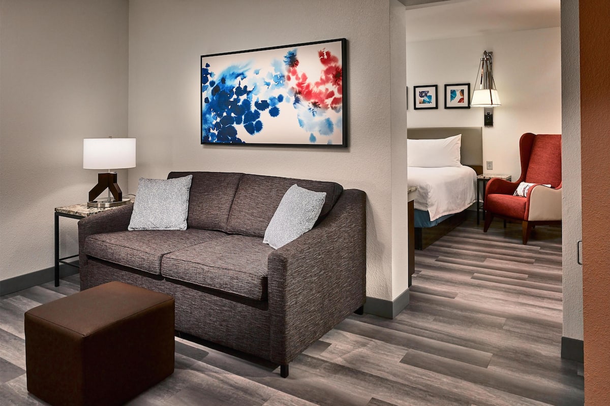 A modern hotel room with a grey sofa, a small table with a lamp, a red armchair, a bed with white bedding, and a large abstract painting on the wall.