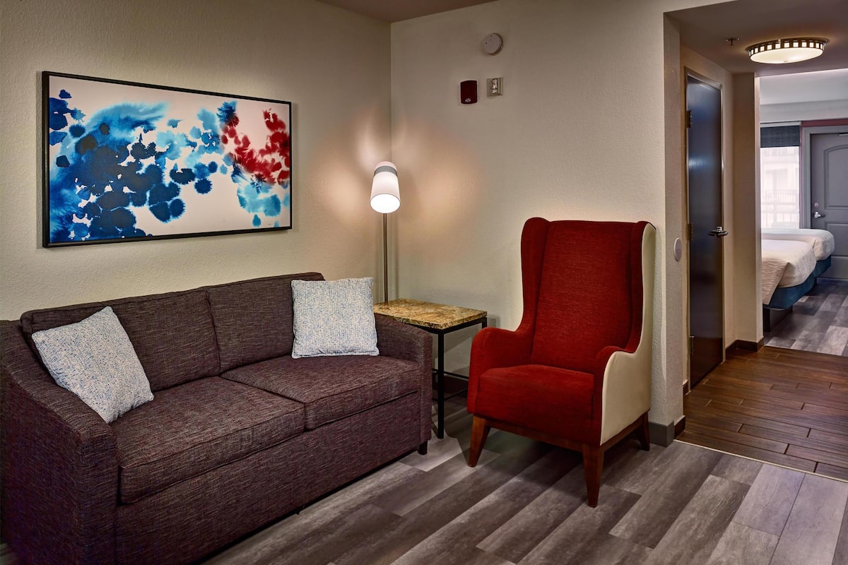 A hotel room with a brown sofa, a red armchair, a small table, and a painting on the wall.
