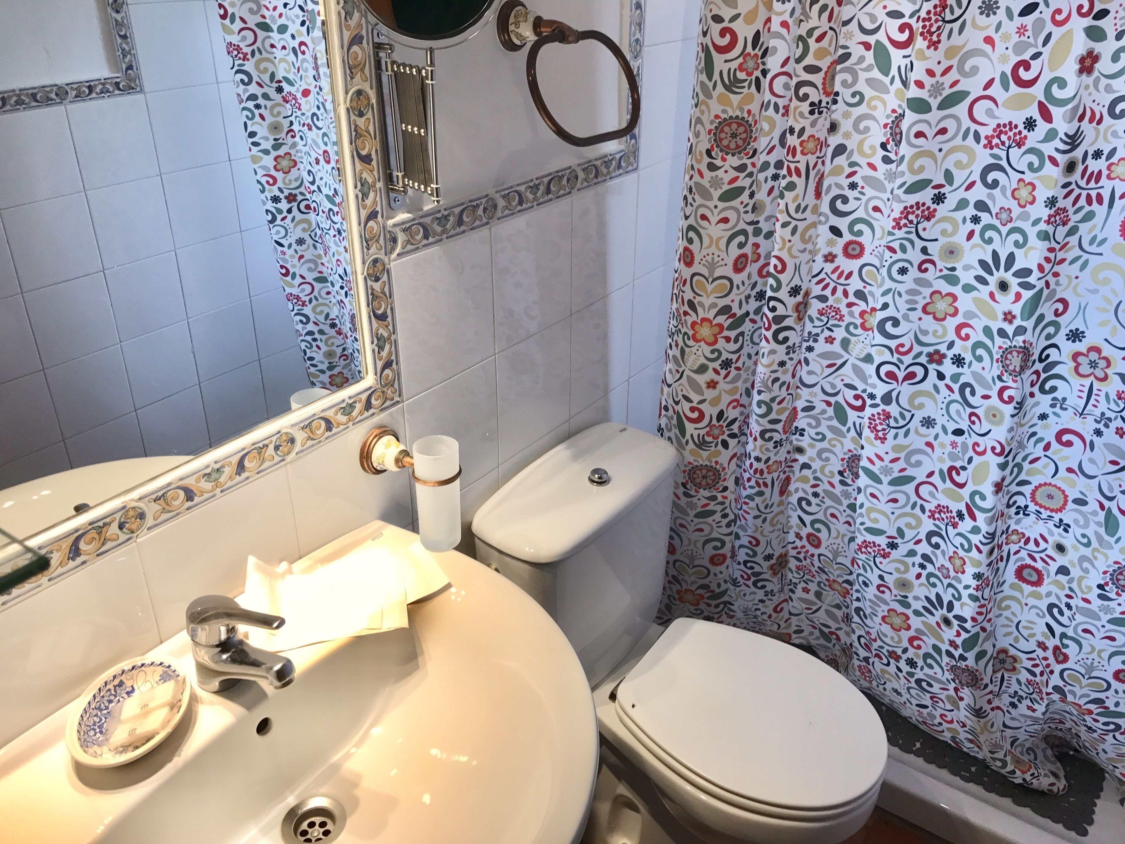 Un baño con lavamanos blanco, inodoro, espejo y cortina de ducha con estampado floral.