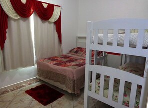 A bedroom with a bunk bed, a single bed with a floral bedspread, a red rug, and a window with red curtains. - Pousada Ares do Sul (Penha)