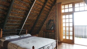 Une chambre avec un plafond en pente, un lit avec un cadre en métal, un mur en briques et une grande fenêtre donnant sur l’extérieur.