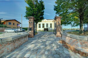 Una entrada pavimentada con muros de ladrillo y una reja que da a un edificio de fachada clara y un ventanal grande.