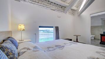 A bedroom with a large bed, a bedside lamp, a view of the sea through a window, and a small table.