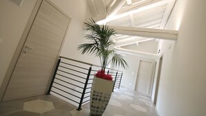 A hallway with a modern design, featuring a geometric patterned floor, a metal railing, and a potted plant with red flowers. - Resilienza (Mira)