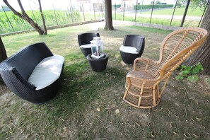 Un área de estar en un jardín, con sillas de mimbre y una mesa, rodeada de césped y árboles.