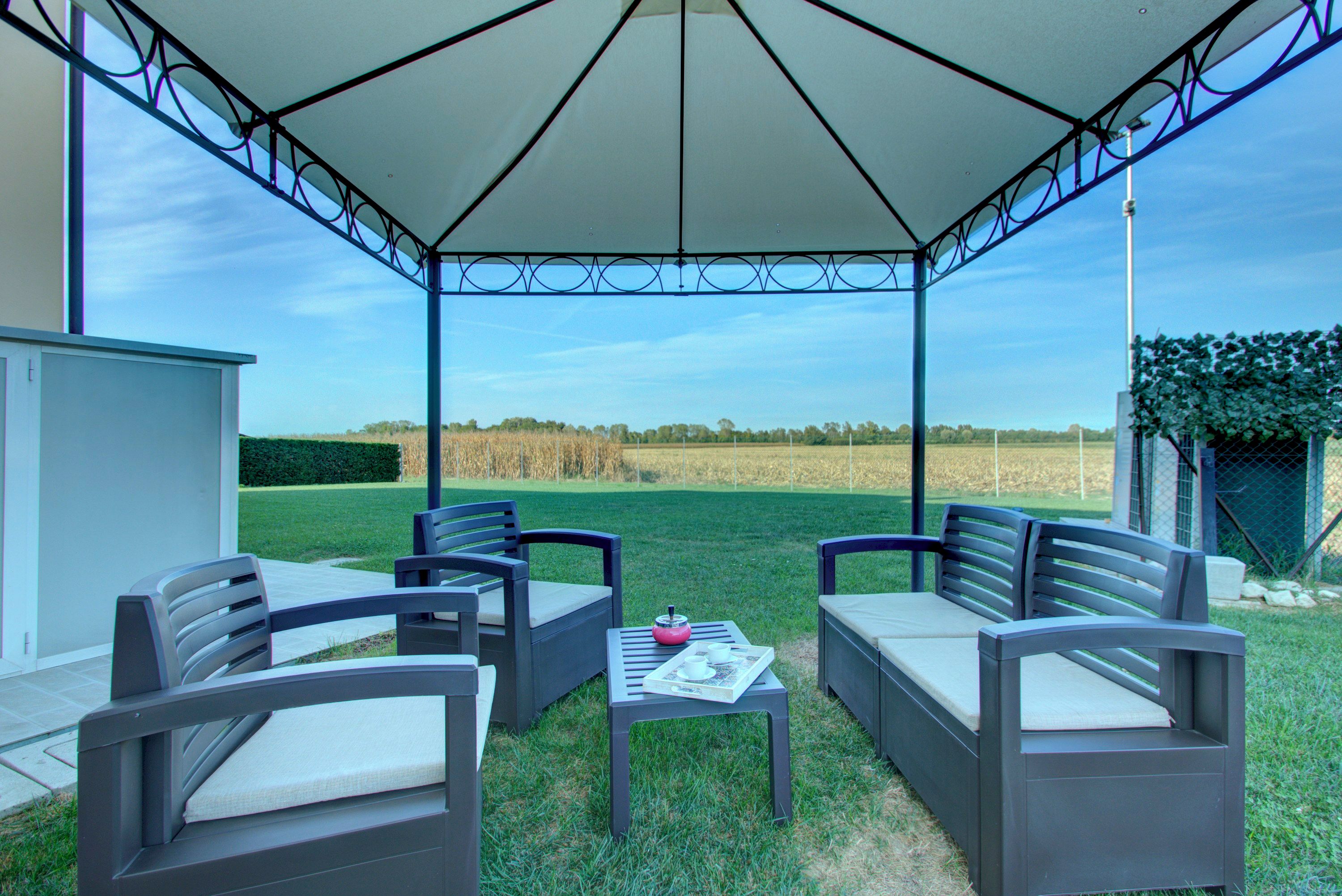 Patio esterno con due divani grigi e un tavolo, un ombrellone bianco e vista su un campo.