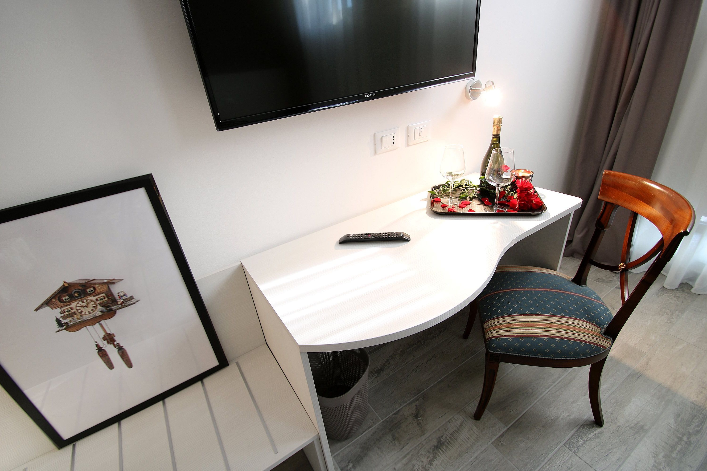 Une chambre d’hôtel dotée d’un téléviseur à écran plat fixé au mur, d’un bureau blanc avec une télécommande, d’une horloge à coucou encadrée, d’une chaise en bois avec un coussin à rayures et d’une petite table sur laquelle se trouvent une bouteille, un verre et des fleurs.