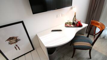Une chambre d’hôtel dotée d’un téléviseur à écran plat fixé au mur, d’un bureau blanc avec une télécommande, d’une horloge à coucou encadrée, d’une chaise en bois avec un coussin à rayures et d’une petite table sur laquelle se trouvent une bouteille, un verre et des fleurs.