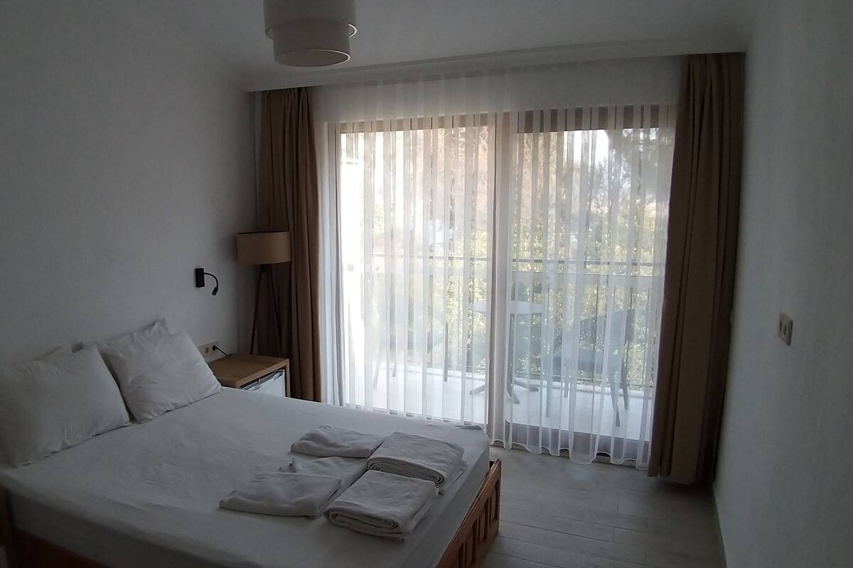 A hotel room with a large bed, a bedside table, a lamp, and a view through sheer curtains to the outside.