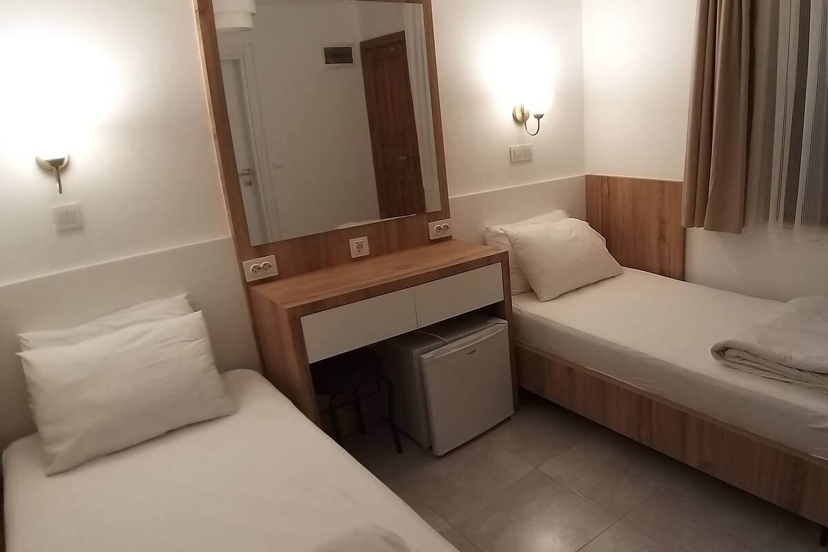 A compact hotel room with two single beds, a small desk, a refrigerator, and a mirror.