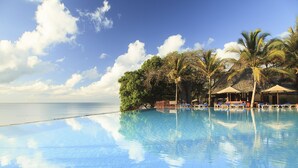 A clear blue infinity pool with lounge chairs and umbrellas, surrounded by palm trees and a view of the ocean.
