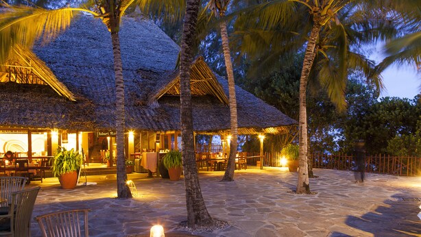 A restaurant with thatched roofs, outdoor seating, and palm trees.